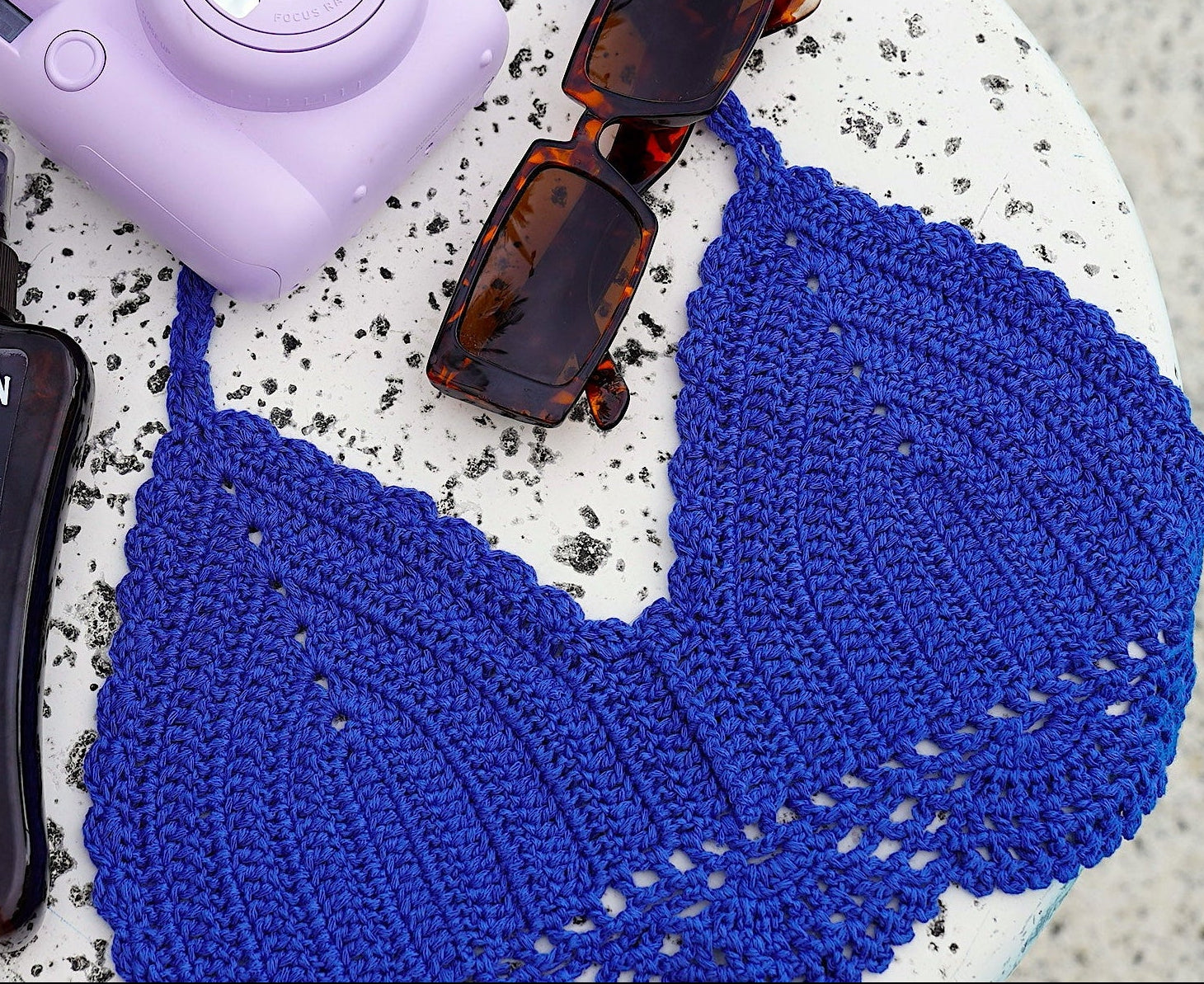 Hawaiian Tropic tanning spray bottle, purple camera, and sunglasses on a round stone surface with blue woven mat.